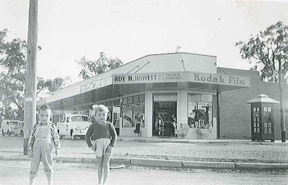Black and white image of 2 small boys standing in the road in front of a series of shops on the corner of Martindale & Pegus Streets in Thornlie.  A sign on the fascia identifies the shop on the corner as Roy H Howitt, Thornlie Pharmacy.  A sign advertising Kodak Film is to the right.  There is a phone box to the right of the shops, there are 2 wicker prams and a stroller outside the shop and 3 vehicles parked in the parking area.