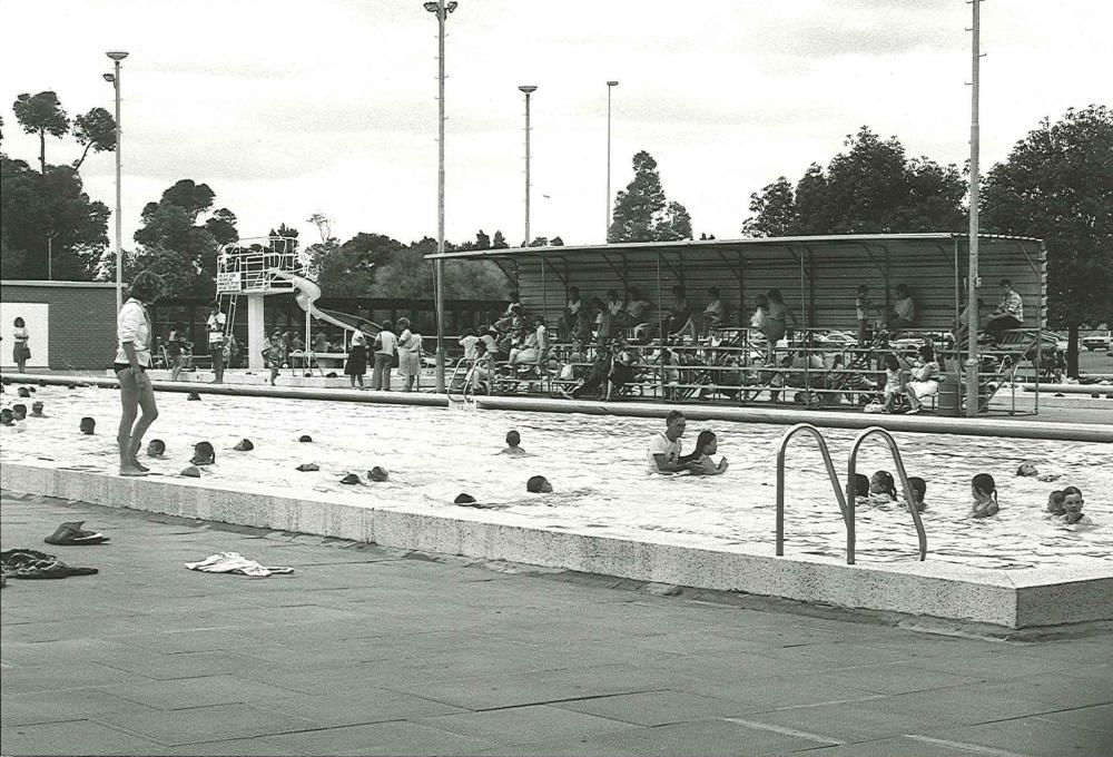 Black and white image of swimming pool with people swimming during swimming lessons and spectators sitting in the stands behind.