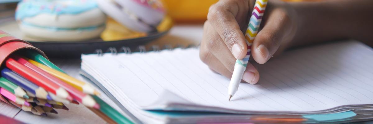 Childs hand holding pen over a lined notebook, pencils and other writing materials strewn on desk