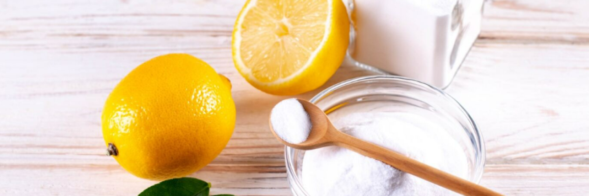 Natural cleaning ingredients, including leaves, lemons, a glass jar of baking soda, a glass bowl of baking soda, and a wooden spoon.