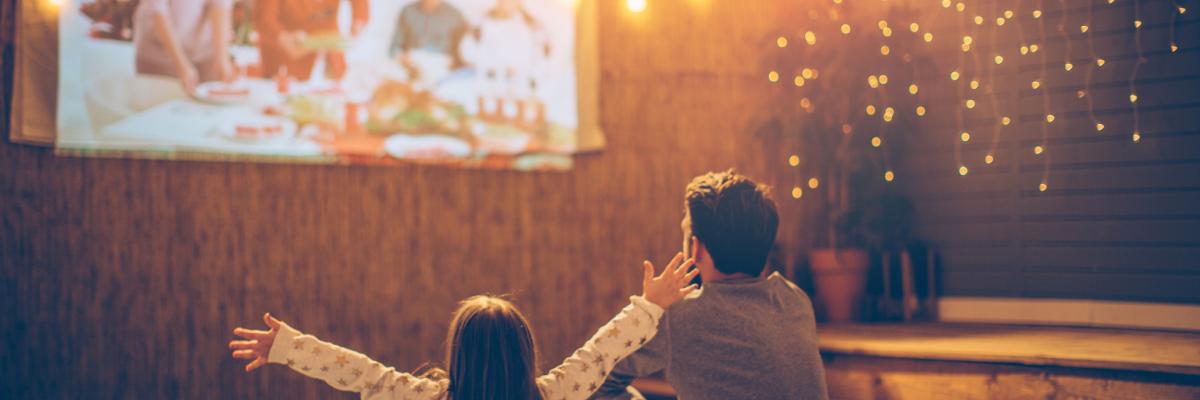 A father and daughter seated in a room lit by fairy lights, looking up at a projector screen on the wall.