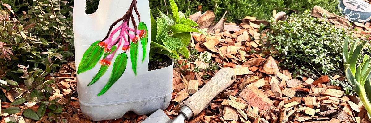 A painted milk bottle that has been upcycled into a miniature greenhouse, sitting on mulch with a trowel nearby