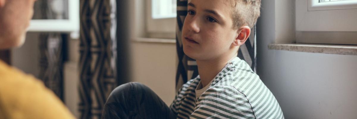 A young pre-teen/teenage boy sitting near a window, with leg drawn up close to him while he looks solemnly at the adult talking to him