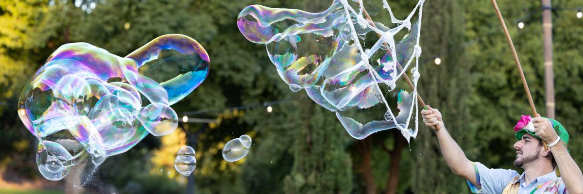 Man dressed up colourfully on the right, blowing big bubbles for kids and adults below to reach