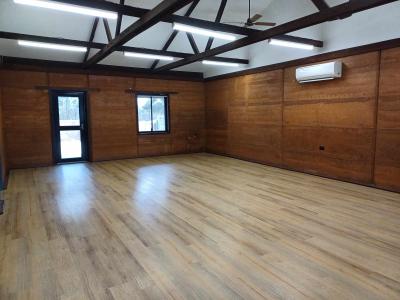Corymbia Room showing front glass door and window, wood-look vinyl flooring, mud brick walls with air conditioner system and wooden roof rafters with fluorescent lights. 