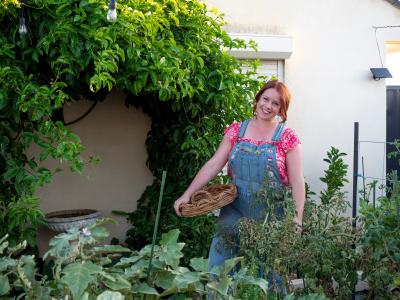 A redheaded woman wearing denim overalls, standing in a lush veggie patch, holding a basket.