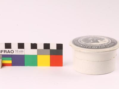 Small white ceramic pot with black printing on lid alongside a standard scale