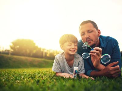 A father and son lying on the grass, with the father blowing bubbles for his smiling child.