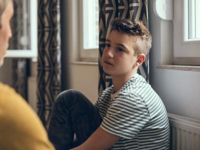 A young pre-teen/teenage boy sitting near a window, with leg drawn up close to him while he looks solemnly at the adult talking to him